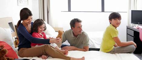 Una familia sentada en una cama en una habitación de hotel, con una televisión y un escritorio.
