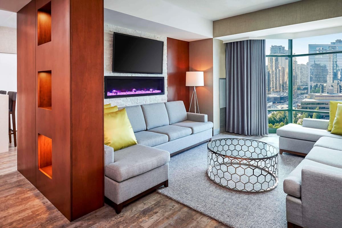 A modern living room with a grey sofa, a fireplace, a TV mounted on a brick wall, and a city view through large windows.