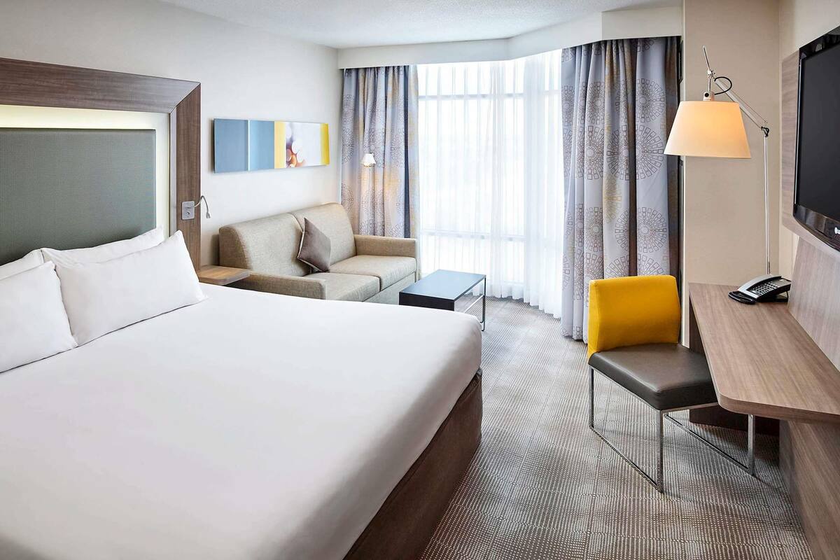 A hotel room with a large bed, a desk with a lamp, a chair, a sofa, and a TV.