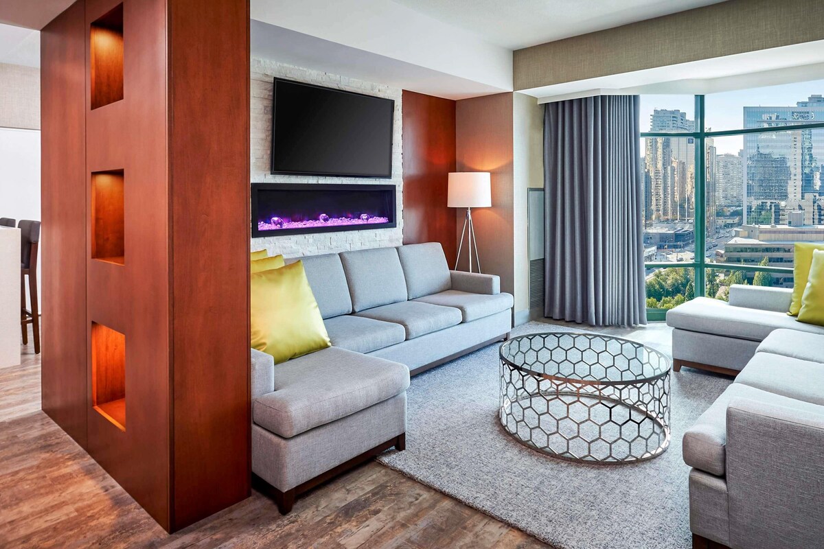 A modern living room with a grey sofa, a fireplace, a TV mounted on a brick wall, and a city view through large windows.
