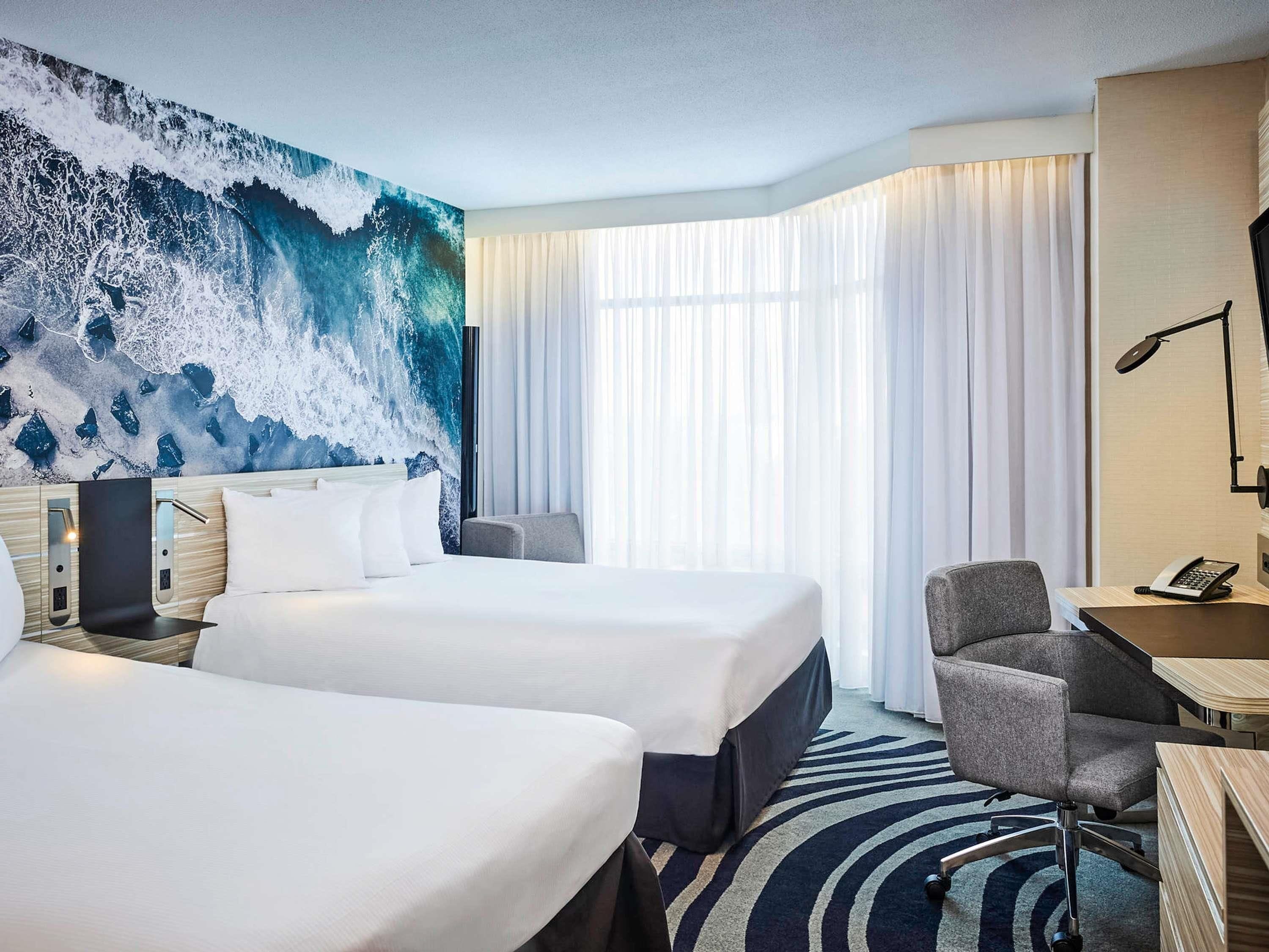 A hotel room with two beds, a desk with a chair, a large window with curtains, and a wall mural.