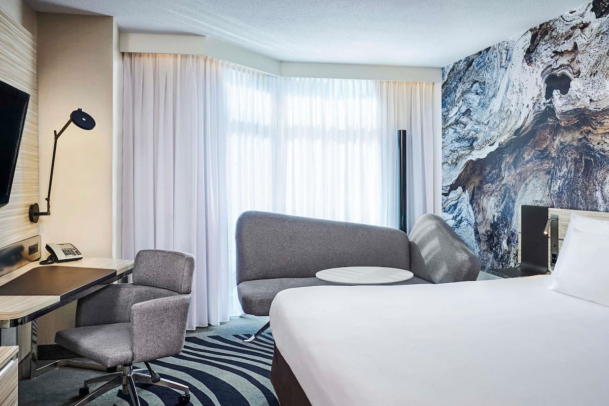 A hotel room with a bed, two armchairs, a desk with a telephone, a TV, and a large window with curtains.