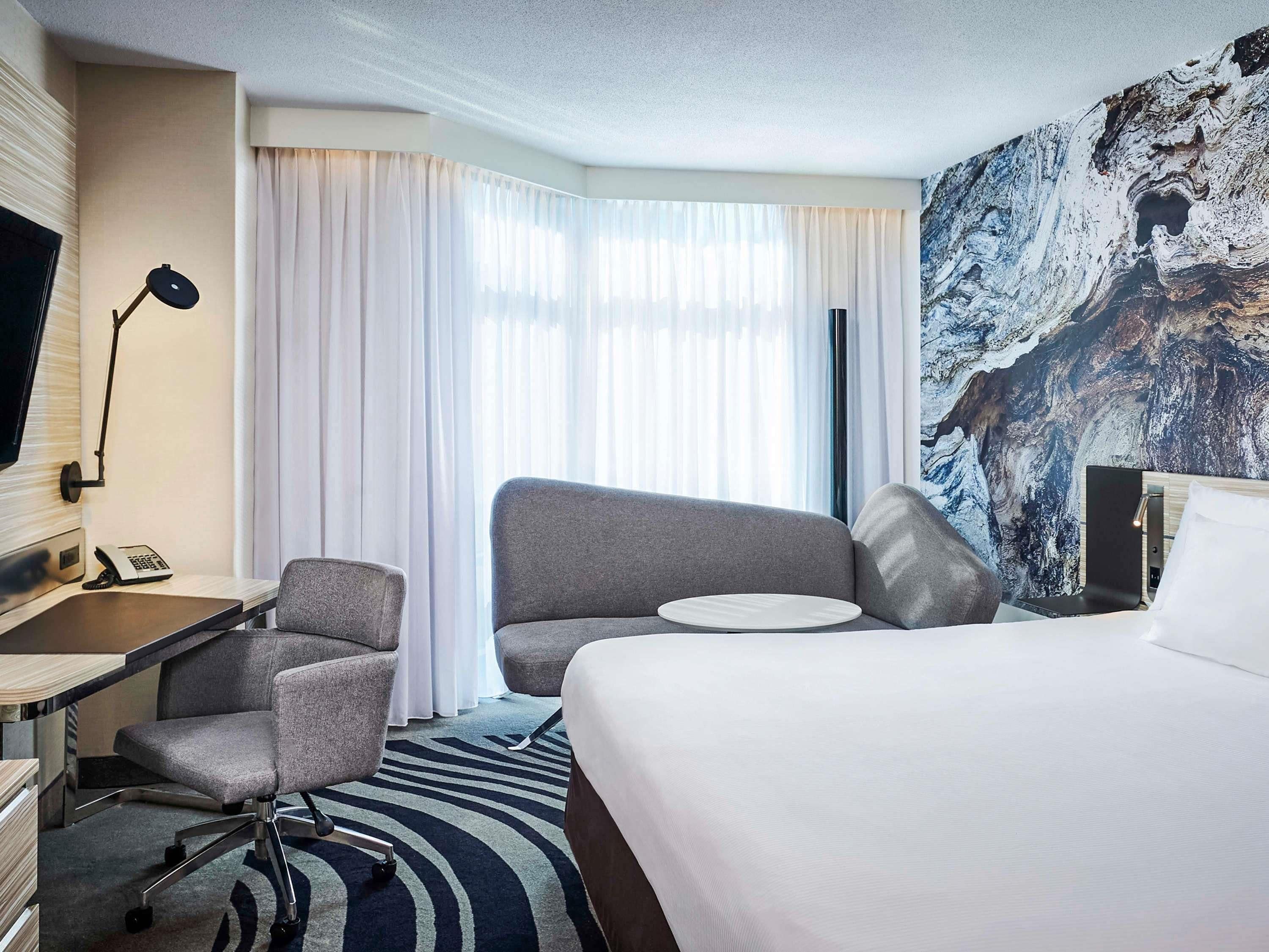 Een hotelkamer met een bed, twee fauteuils, een bureau met telefoon, een televisie en een groot raam met gordijnen.