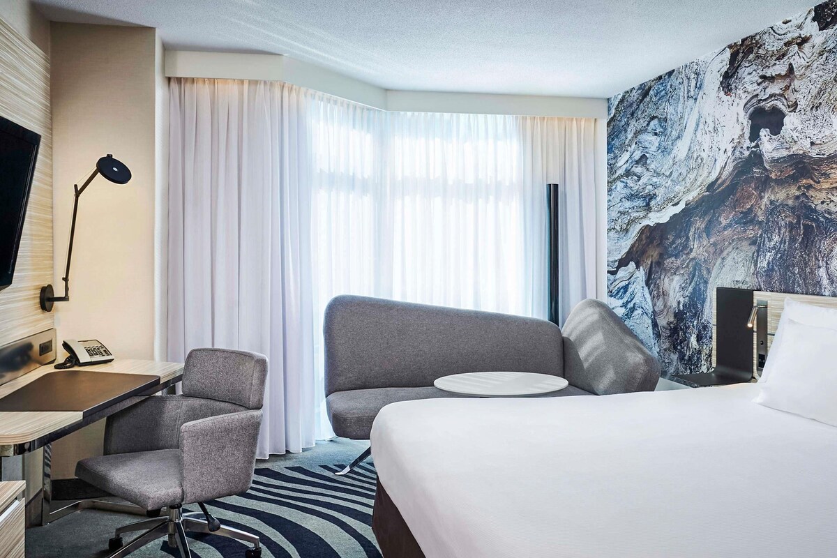 A hotel room with a bed, two armchairs, a desk with a telephone, a TV, and a large window with curtains.