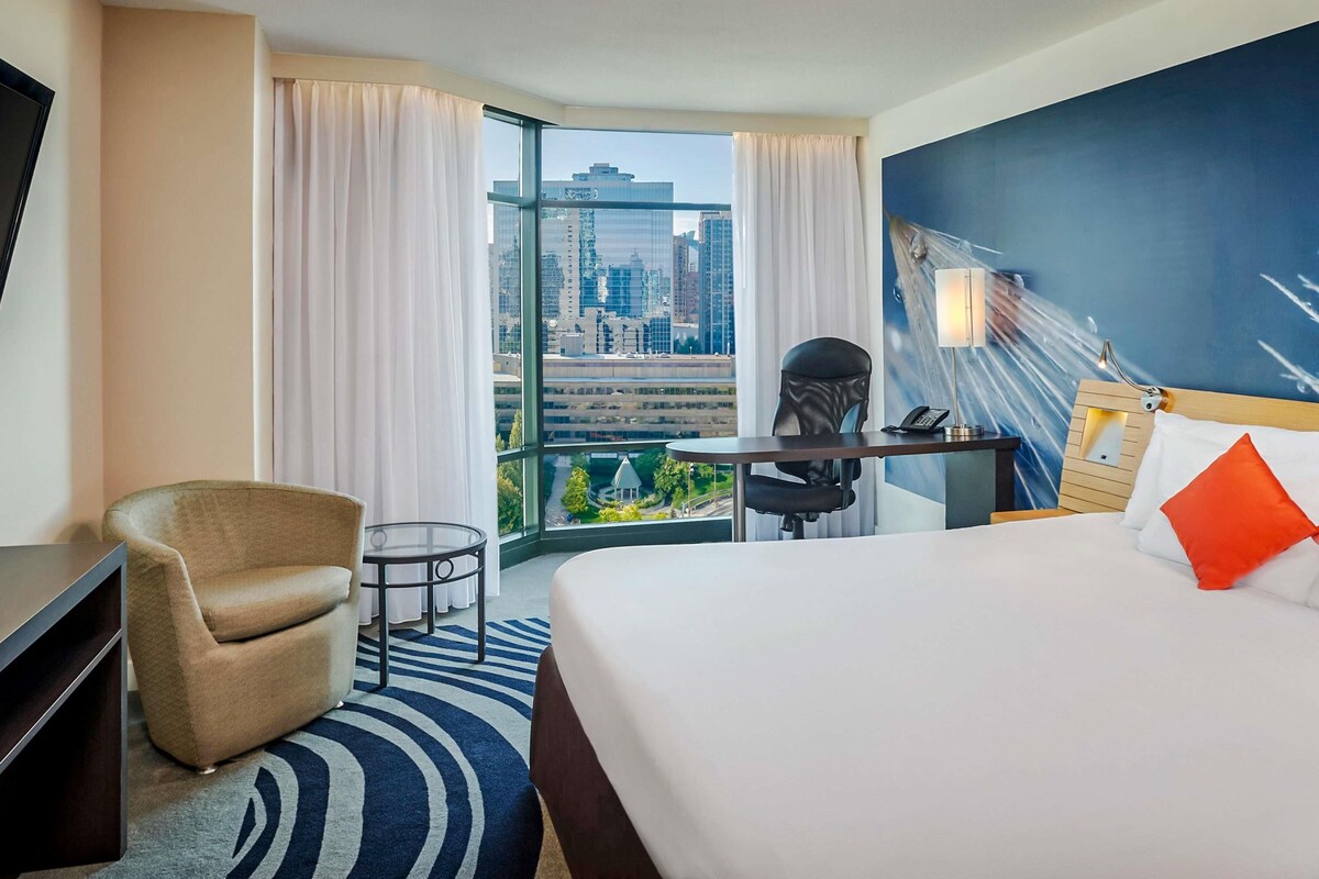A hotel room with a bed, a desk, a chair, a TV, and a view of a cityscape through large windows.