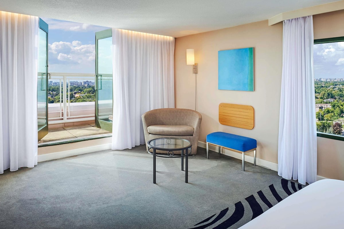 A hotel room with a large window, a chair, a small table, a blue bench, and a painting on the wall.