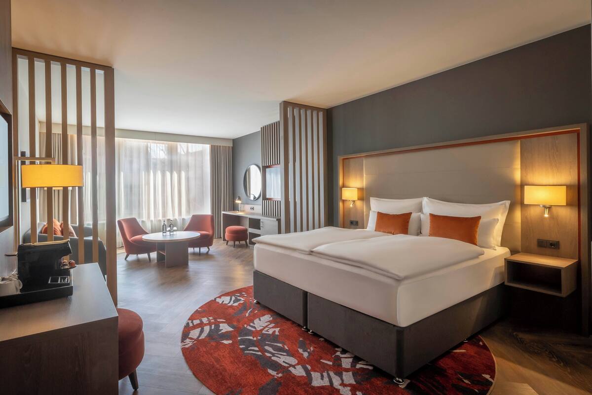 Habitación de hotel con una cama grande, un escritorio con lámpara, una silla, una mesita pequeña y un ventanal con cortinas.