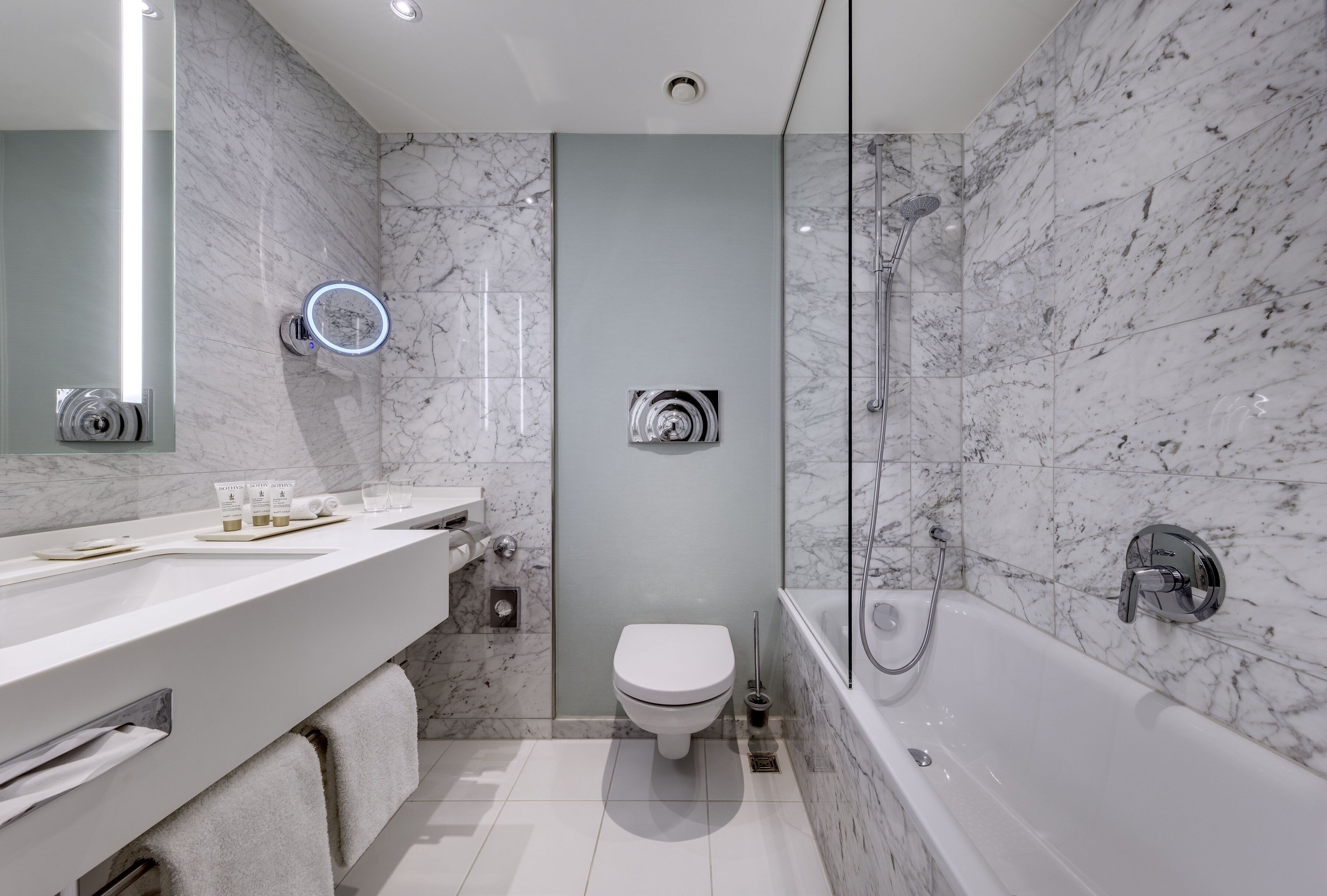 A modern bathroom with a marble wall, a white toilet, a bathtub, and a vanity with a mirror and lights.