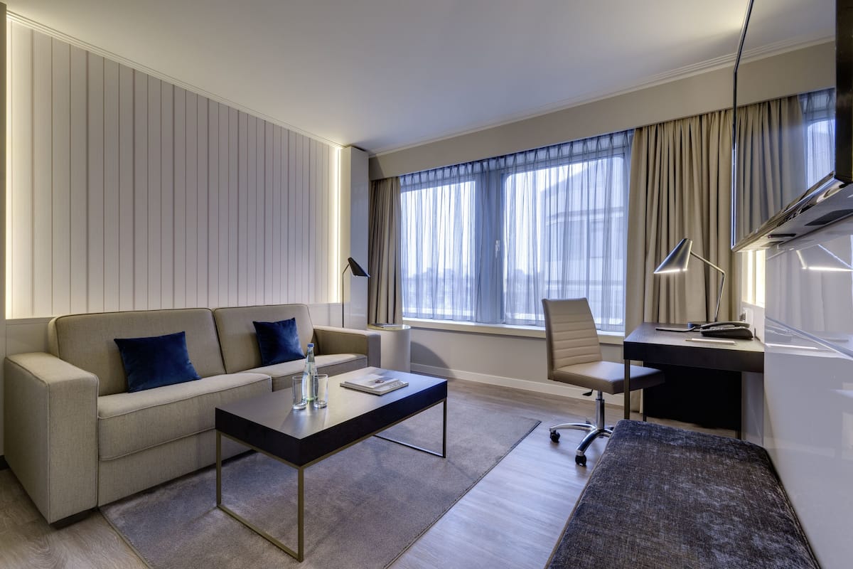 Una habitación de hotel moderna con sofá, escritorio, silla y un gran ventanal con cortinas.