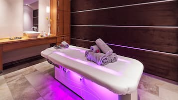 Relax in a serene spa treatment room with a central massage table, a large mirror, and a vanity with sink and toiletries.