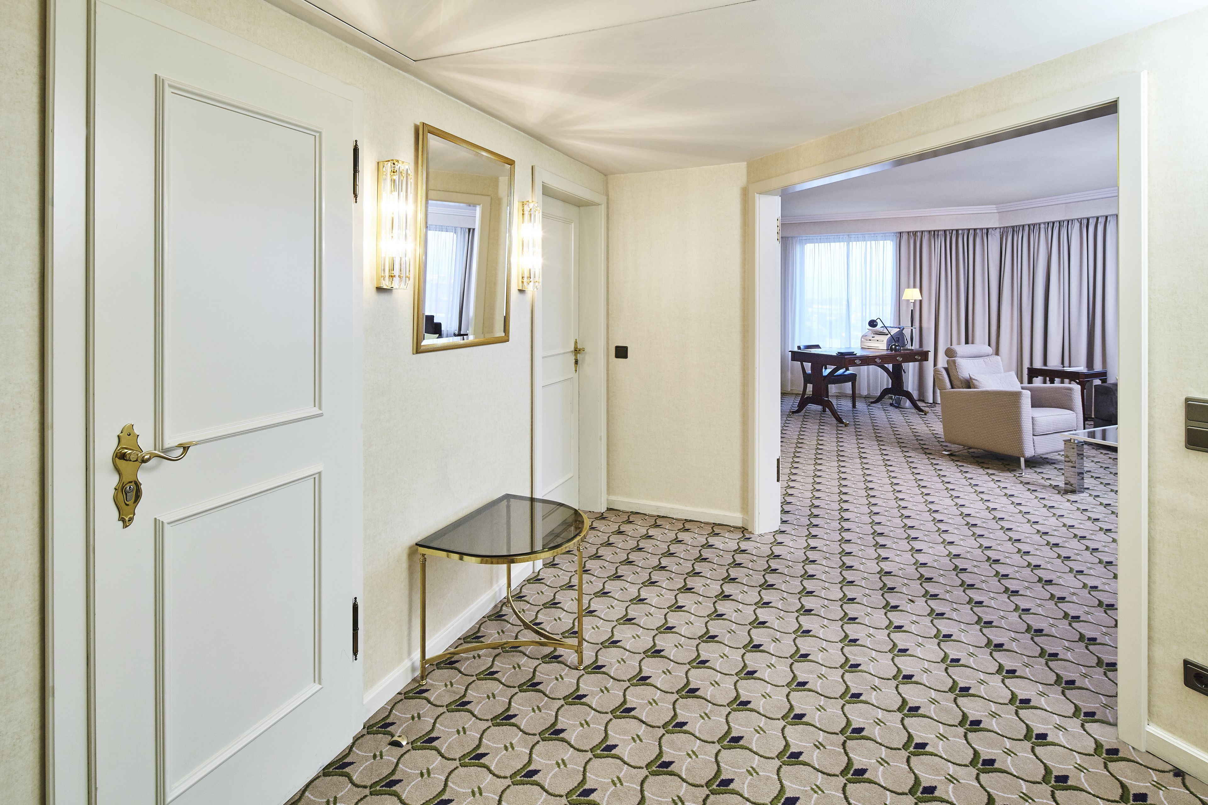 Un corridoio di un hotel con una moquette fantasia, un tavolino con piano in vetro, uno specchio e una zona pranzo con tavolo e sedie.