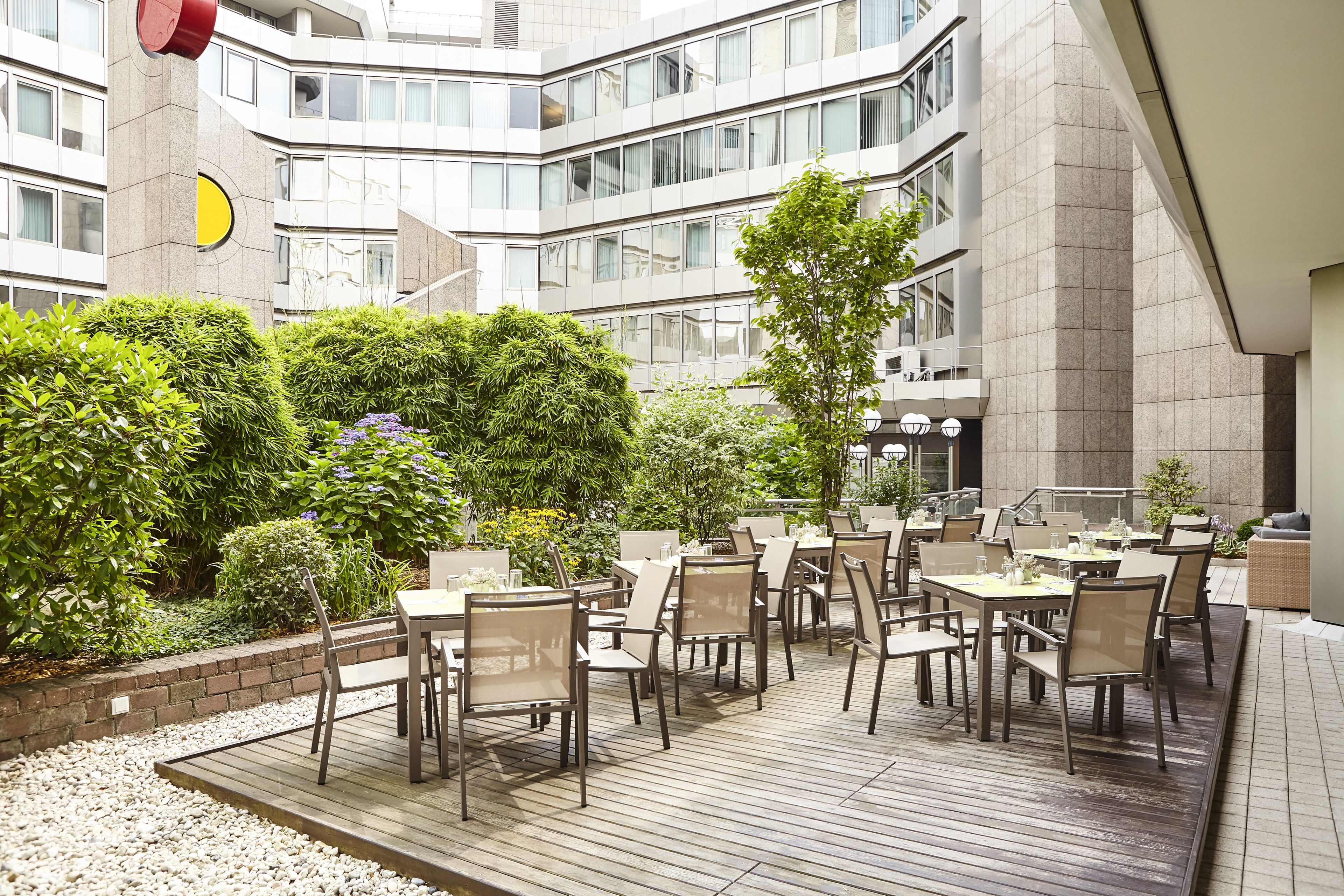A courtyard with outdoor seating, surrounded by greenery and a modern building facade.
