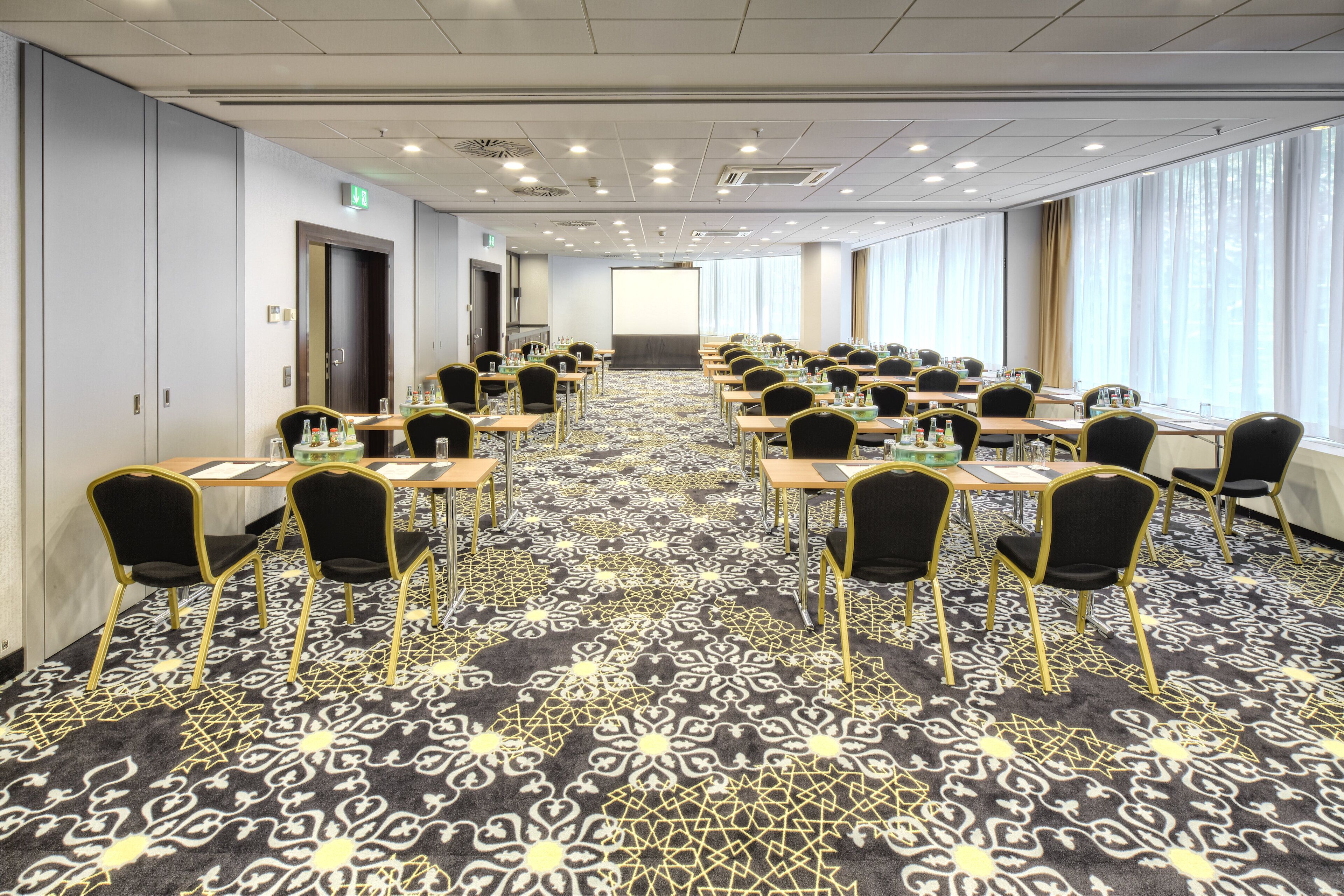 Un salón de conferencias con filas de mesas y sillas, una alfombra estampada y ventanas amplias con cortinas.