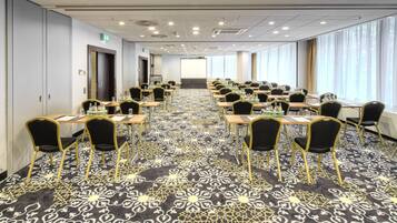 Un salón de conferencias con filas de mesas y sillas, una alfombra estampada y ventanas amplias con cortinas.