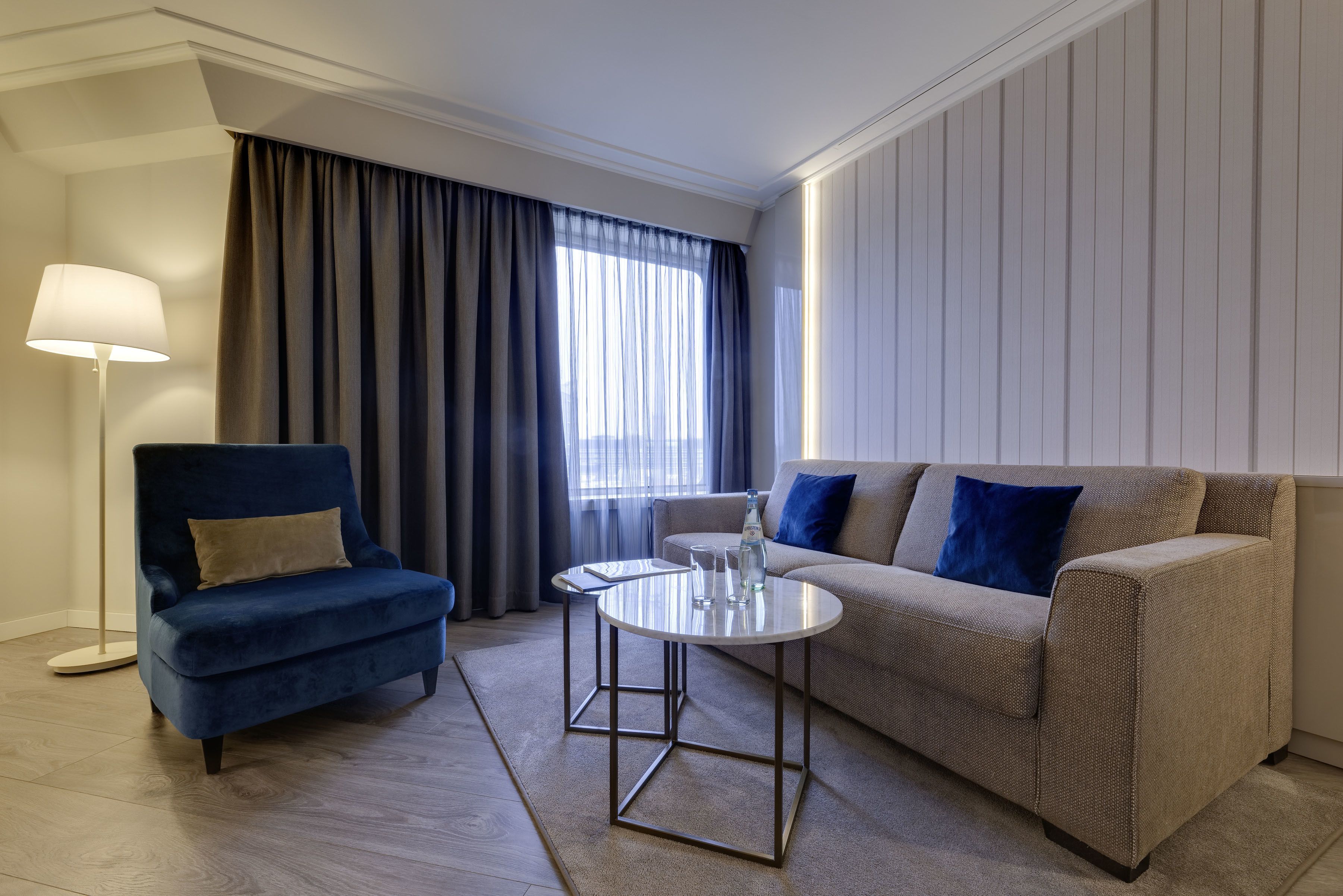 A modern living room with a blue armchair, a beige sofa, a round coffee table, and a floor lamp.