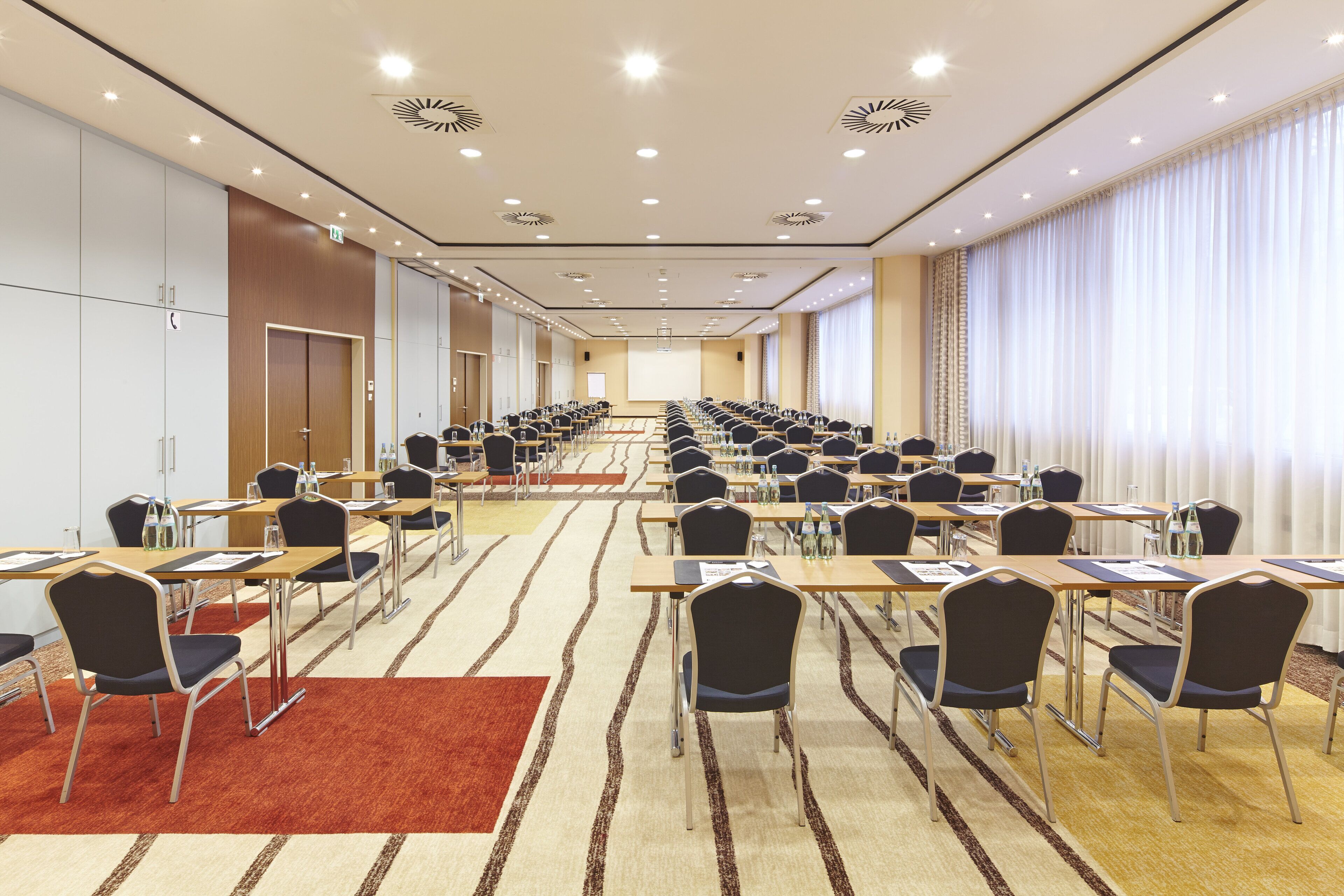 A conference room with rows of tables and chairs, a projector screen, and a large window with curtains.