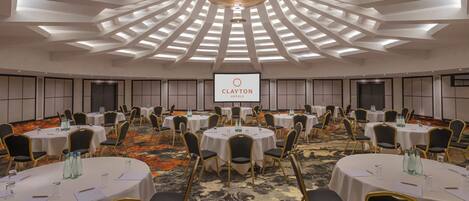 Un salón de conferencias con varias mesas y sillas, una pantalla grande mostrando el logotipo de Clayton Hotels y un diseño de techo moderno.