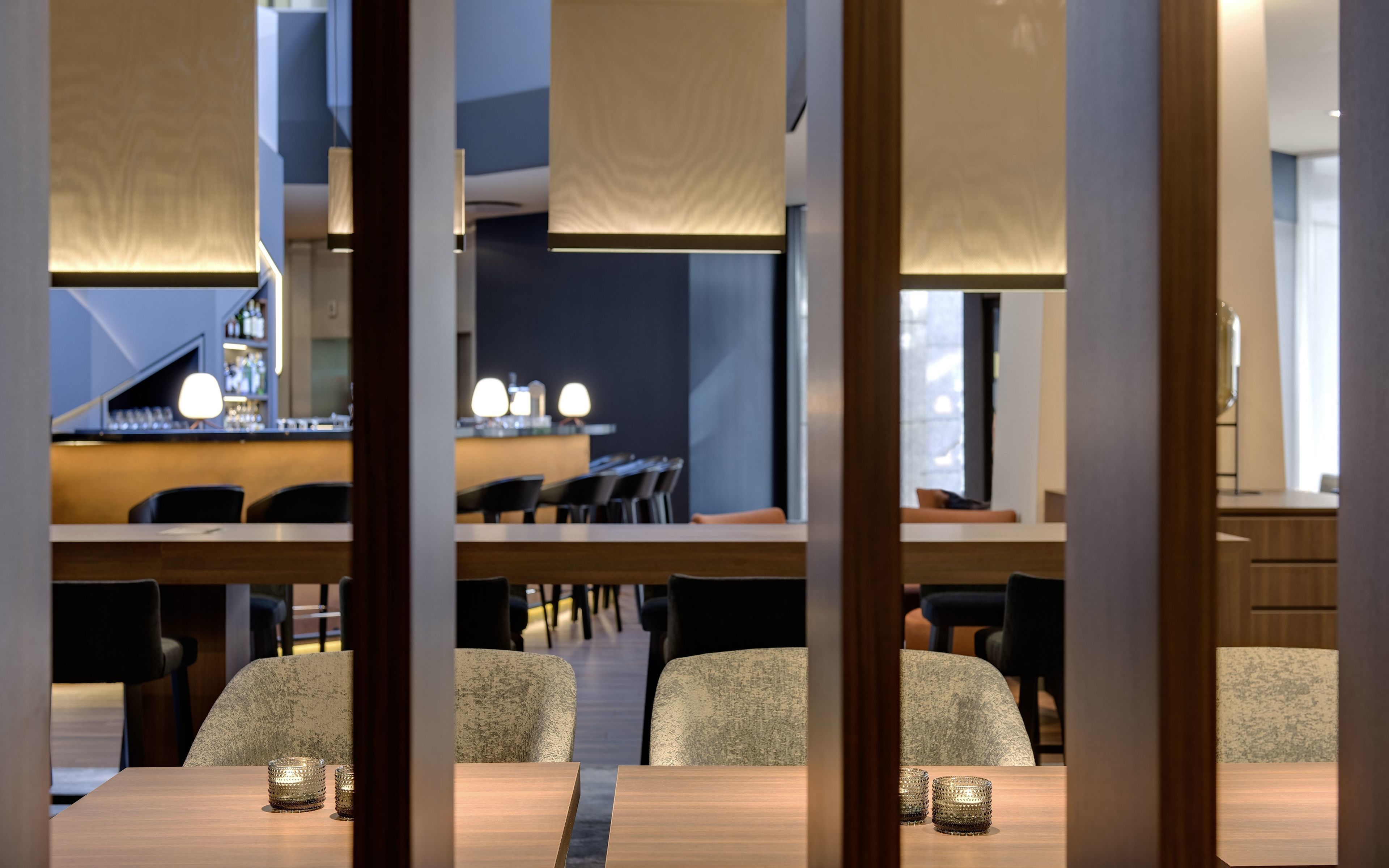 Un área de comedor moderna con mesas y sillas de madera, un bar al fondo e iluminación colgante.