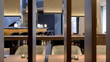 Un área de comedor moderna con mesas y sillas de madera, un bar al fondo e iluminación colgante.