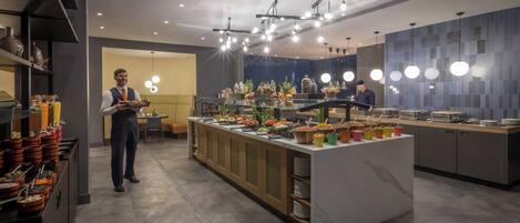 Un área de bufé moderna en un hotel, con diversas opciones de comida, un espacio para sentarse y un bar.
