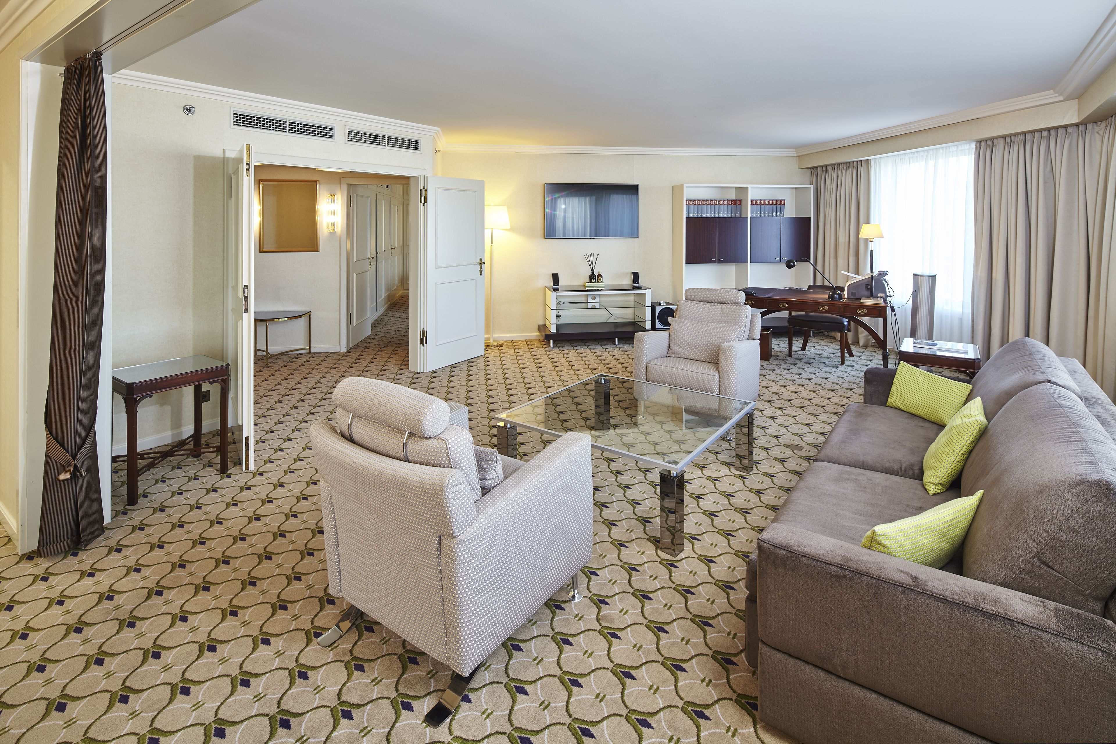 A hotel room with a living area featuring a sofa, armchair, and a glass coffee table. There is a desk with a lamp and a chair, and a bookshelf with books. The room has patterned carpet and a door leading to another room.