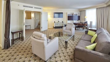 A hotel room with a living area featuring a sofa, armchair, and a glass coffee table. There is a desk with a lamp and a chair, and a bookshelf with books. The room has patterned carpet and a door leading to another room.