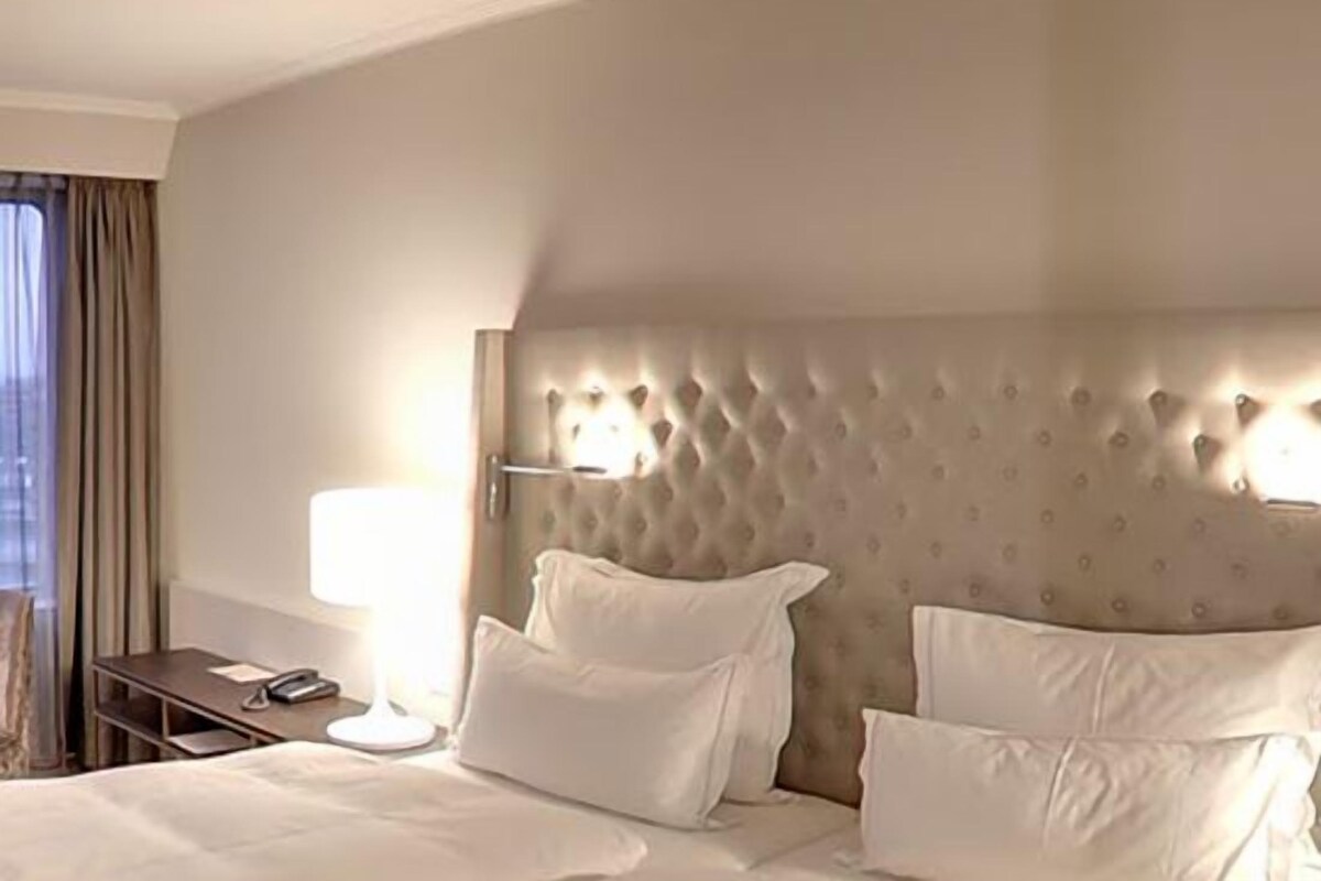 A hotel room with a tufted headboard, a bedside table with a lamp, and a view of the city through the window.