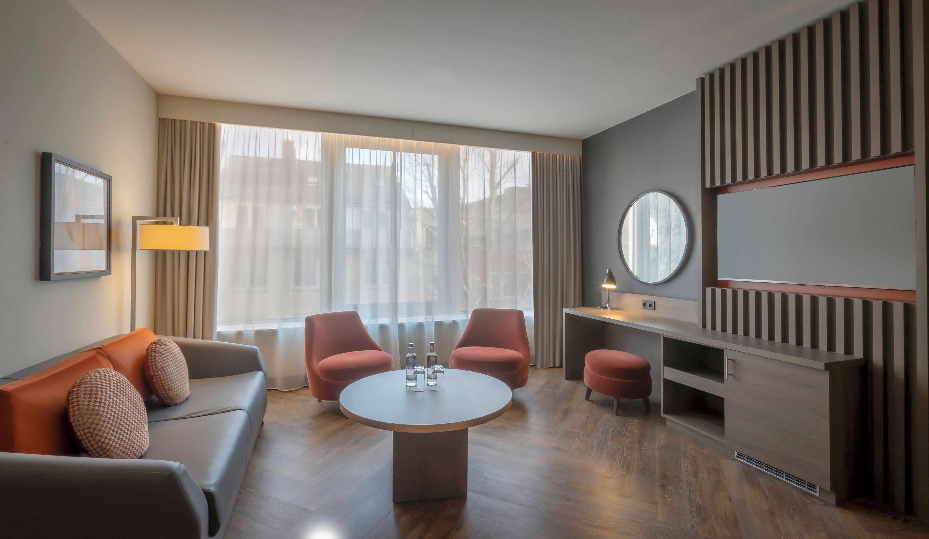 Quarto de hotel moderno com sofá, duas poltronas, uma mesa redonda, escrivaninha, luminária, espelho e janela com cortinas.