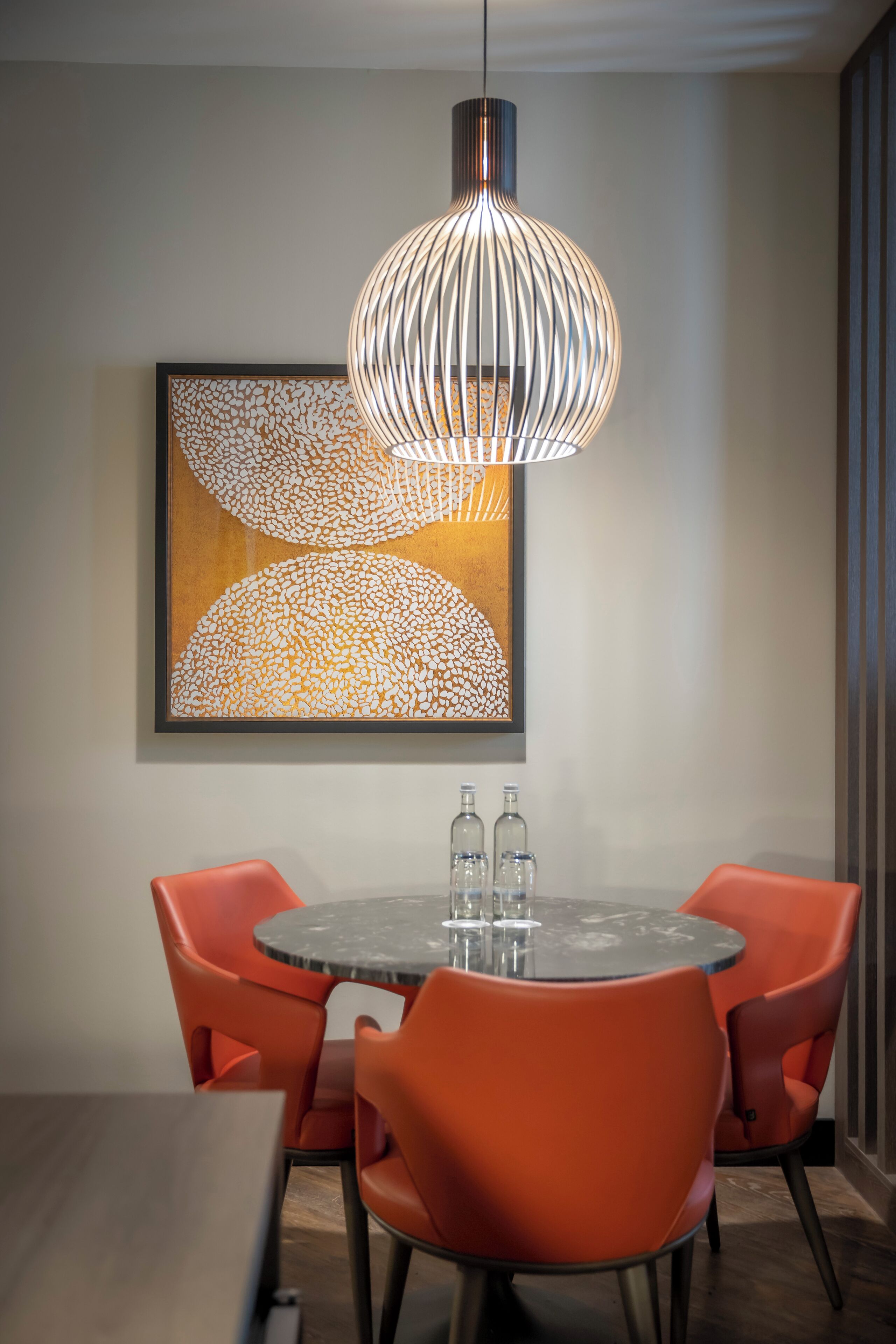 A dining area with a round table, orange chairs, a modern pendant light, and a framed abstract artwork on the wall.