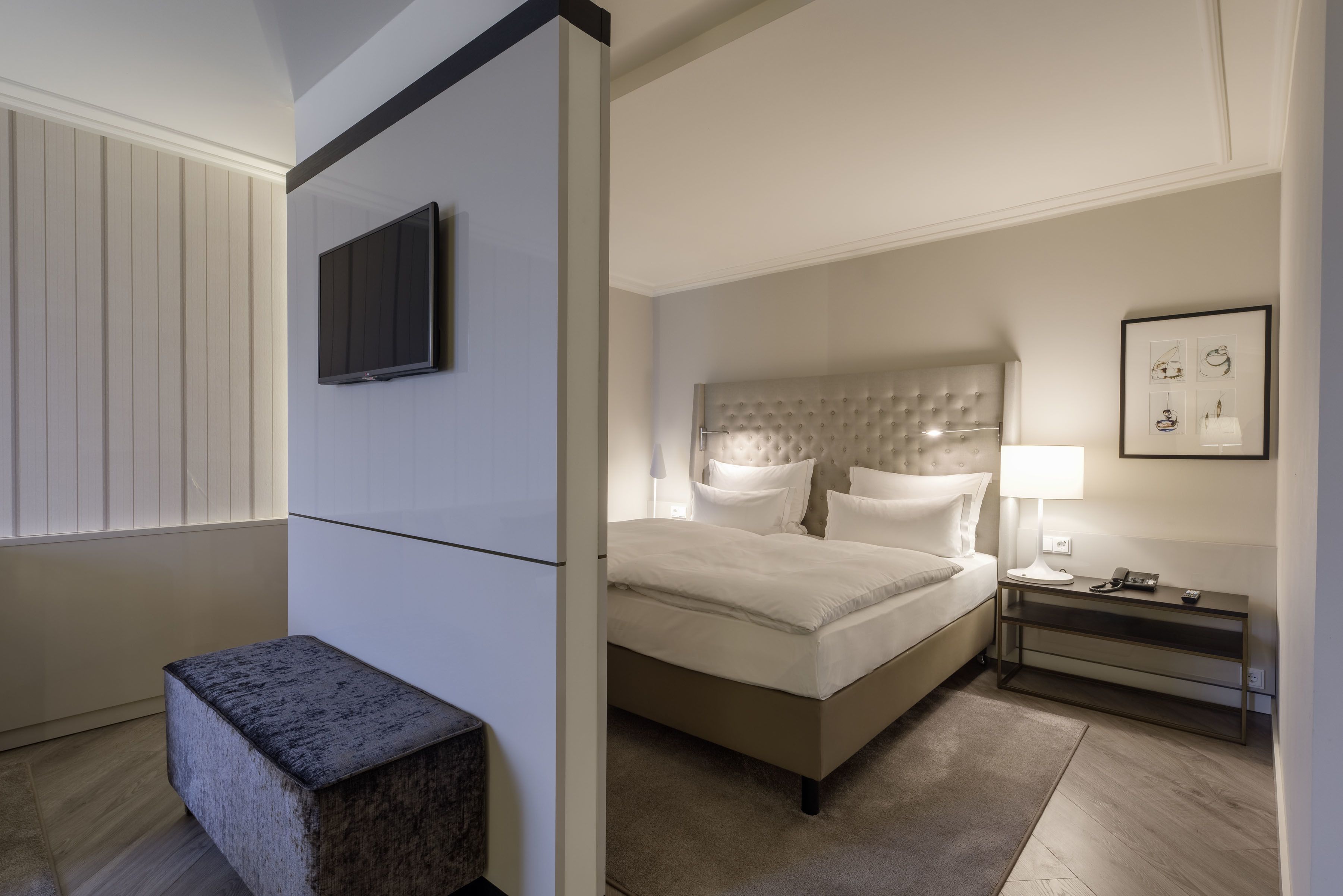 A hotel room with a bed, bedside table, lamp, artwork, and a television.
