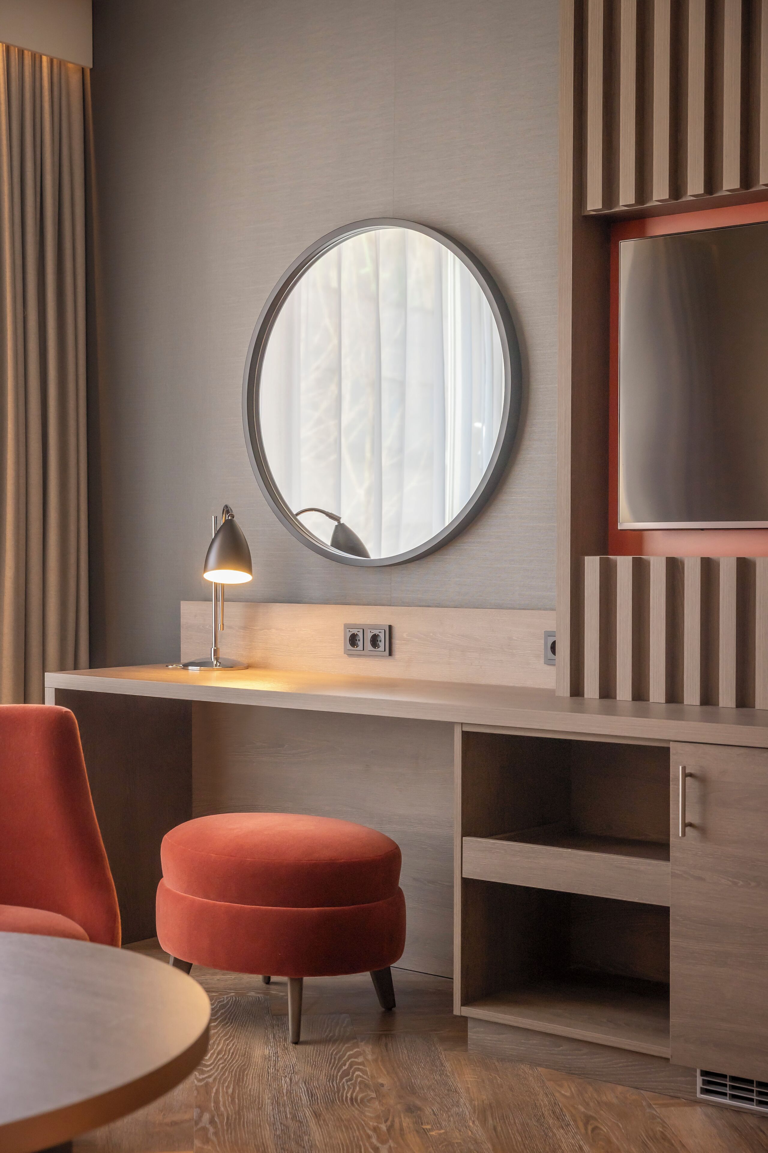 A modern room with a round mirror, a desk with a lamp, a red ottoman, and a TV on a wooden cabinet.