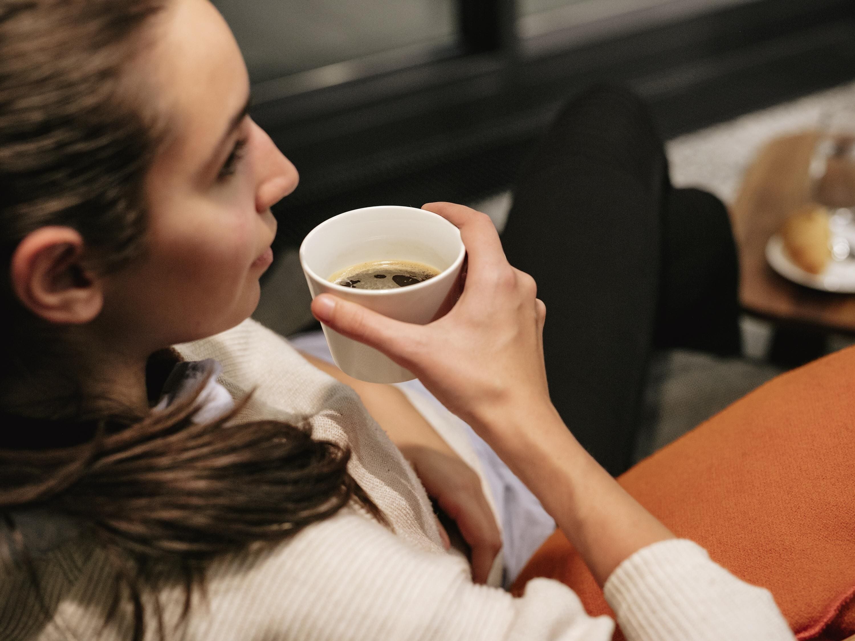 Una persona sosteniendo una taza, probablemente con una bebida, sentada en una habitación con ventana y una mesa con un plato y un vaso.