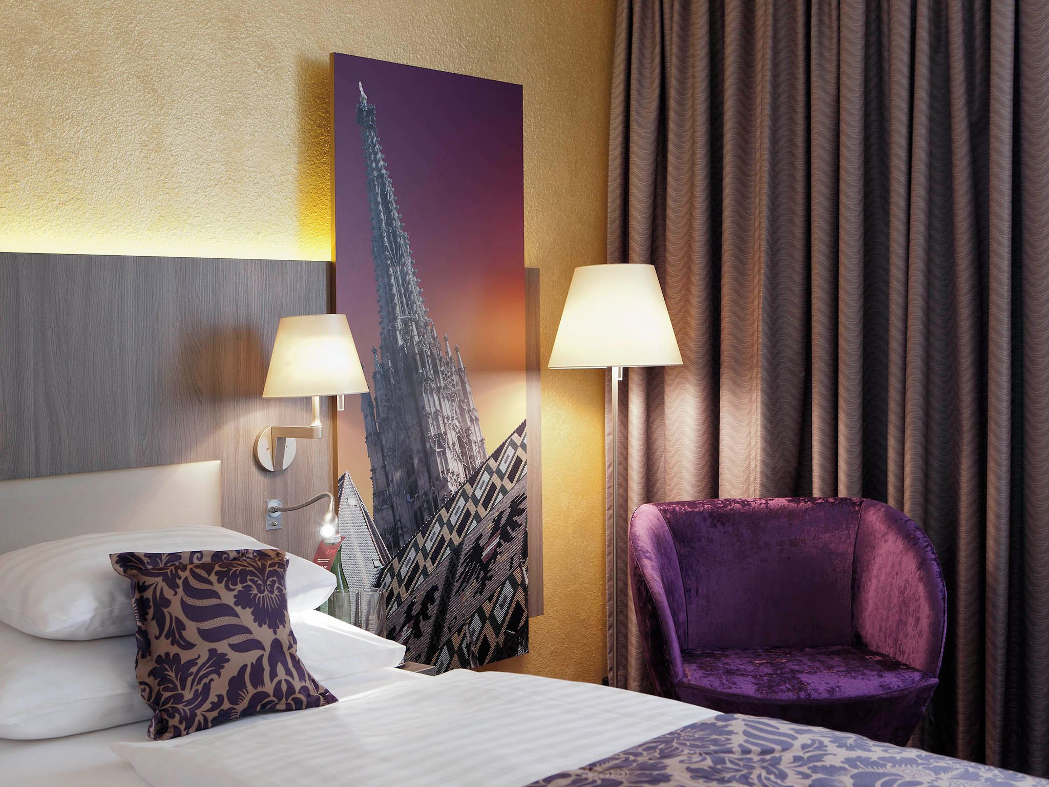 A hotel room with a bed, two lamps, a purple chair, and a large painting of a tower.