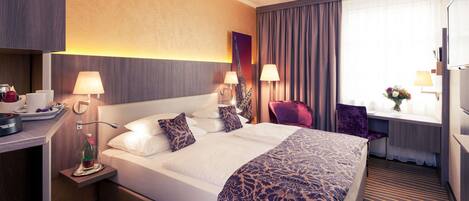 Une chambre d’hôtel avec un grand lit, deux tables de chevet avec des lampes, un bureau avec une chaise, une fenêtre avec des rideaux et une petite table sur laquelle est posé un vase de fleurs.