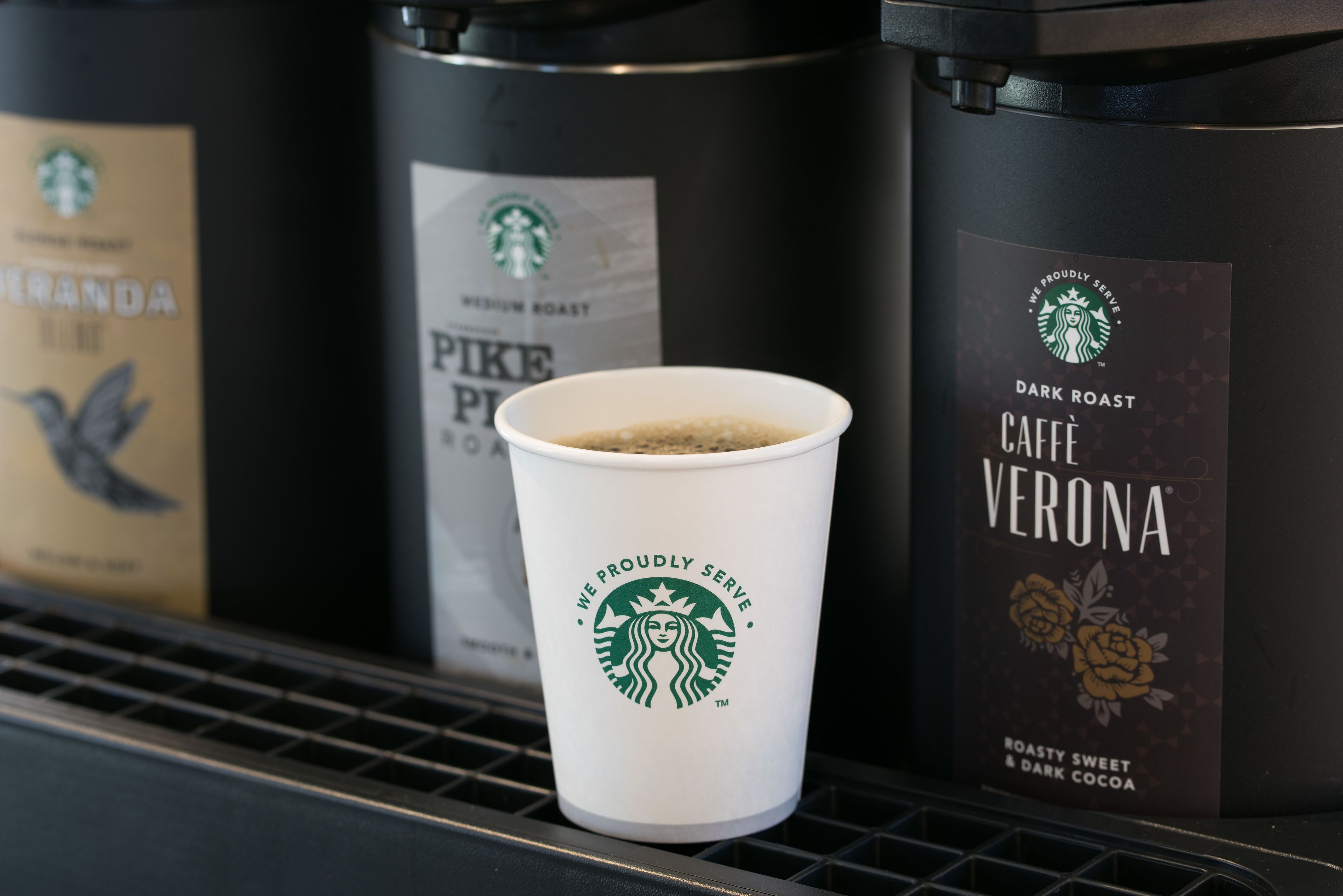 Una taza de Starbucks en un estante con dos cafeteras que dicen Pike Place Roast y Cafe Verona.
