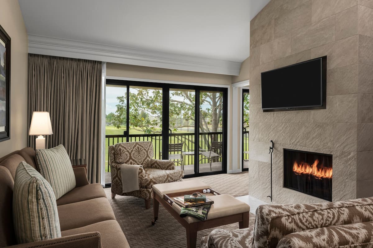 A modern living room with a sofa, armchair, and ottoman, a flat-screen TV, a fireplace, and a balcony with outdoor furniture.