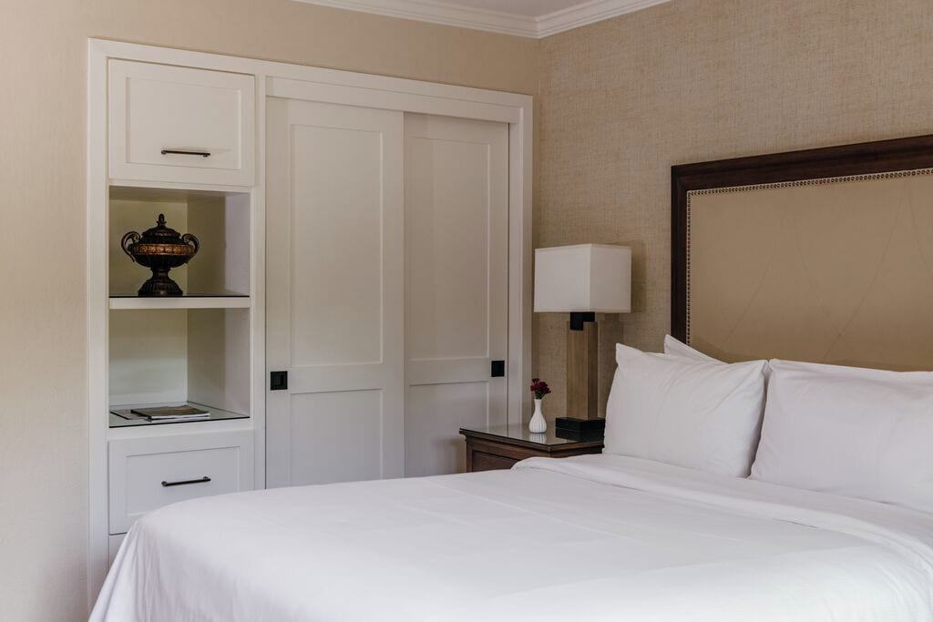A hotel room with a bed, a nightstand, a lamp, a wardrobe, and a shelf with a decorative item.