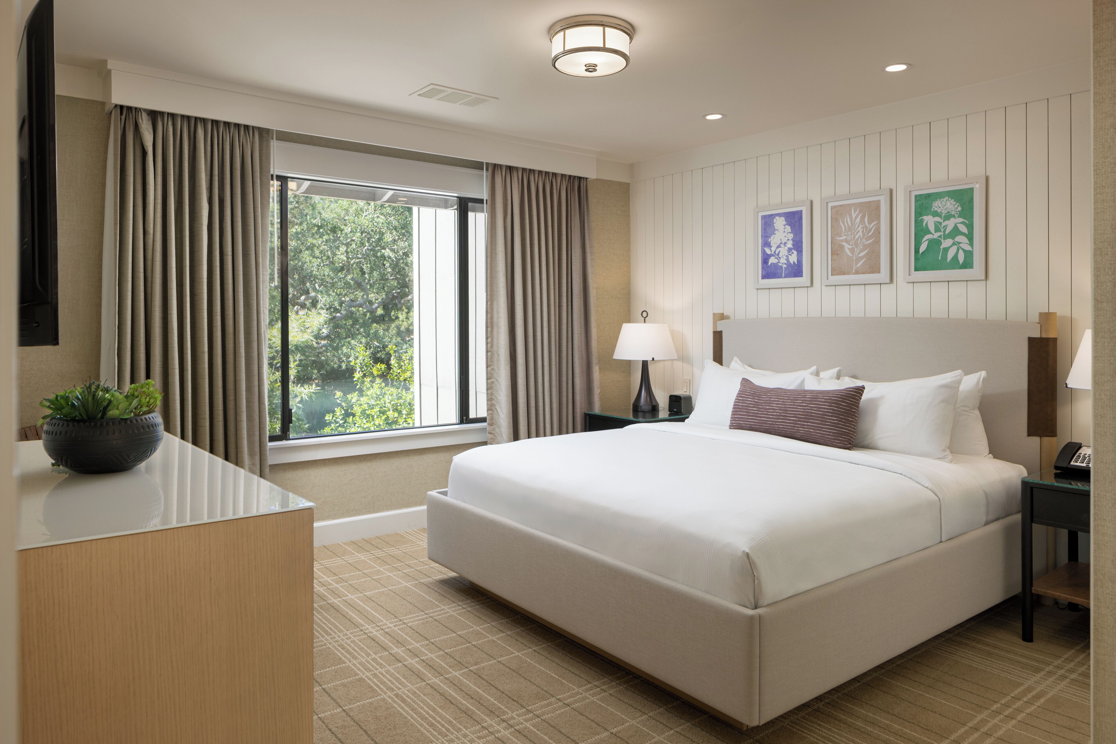 A hotel room with a large bed, a desk with a plant, a window with curtains, and framed artwork on the wall.
