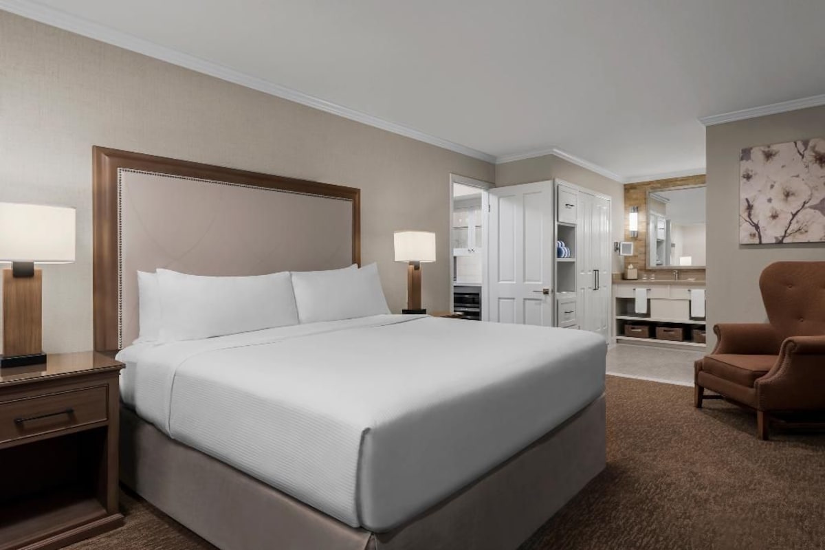 A hotel room with a large bed, bedside tables, a chair, and a bathroom visible through an open door.