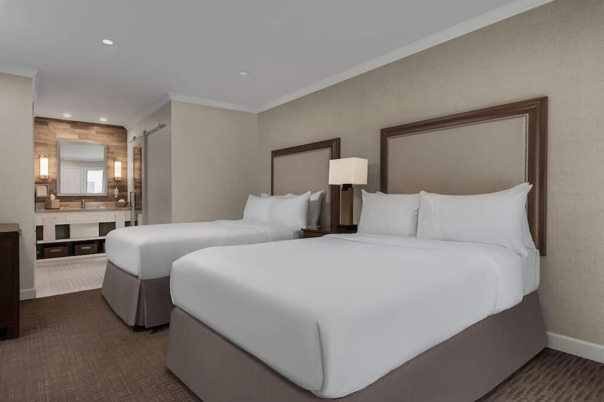 A hotel room with two beds, a large mirror, a vanity with a sink, and a carpeted floor.