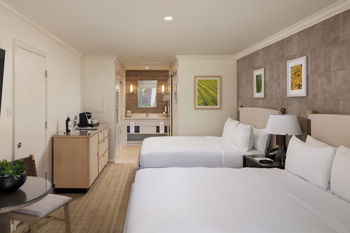 A hotel room with two beds, a desk, a coffee machine, a TV, and a bathroom visible in the background.