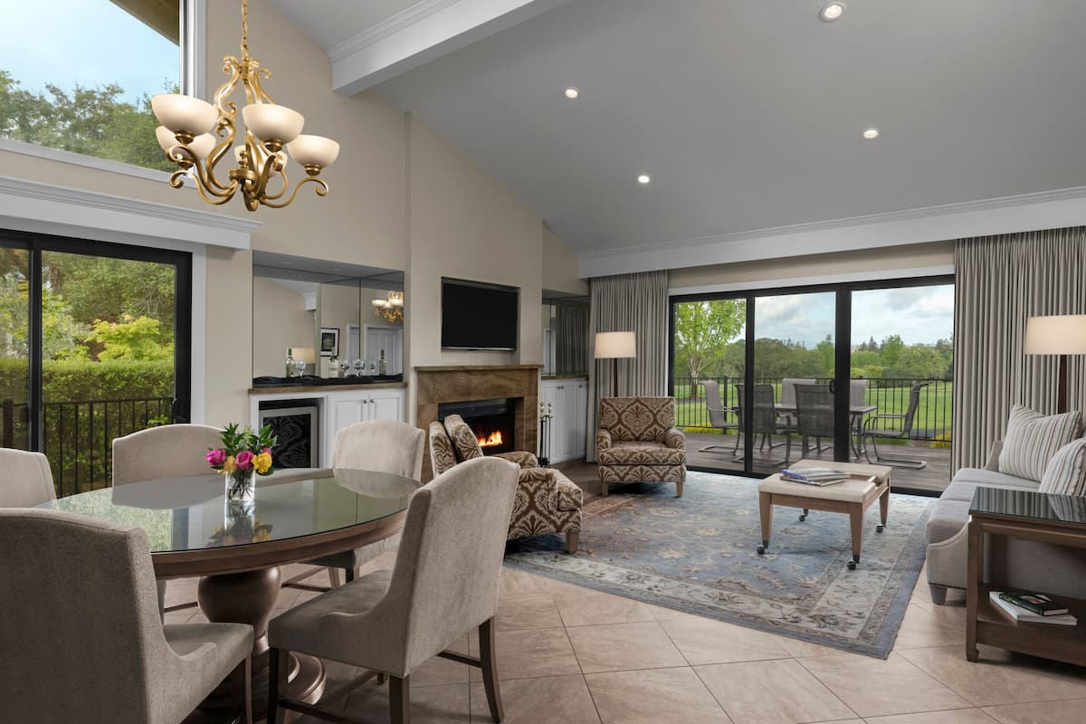 A spacious living room with a glass dining table, comfortable seating, a fireplace, and a large window offering a view of the outdoors.