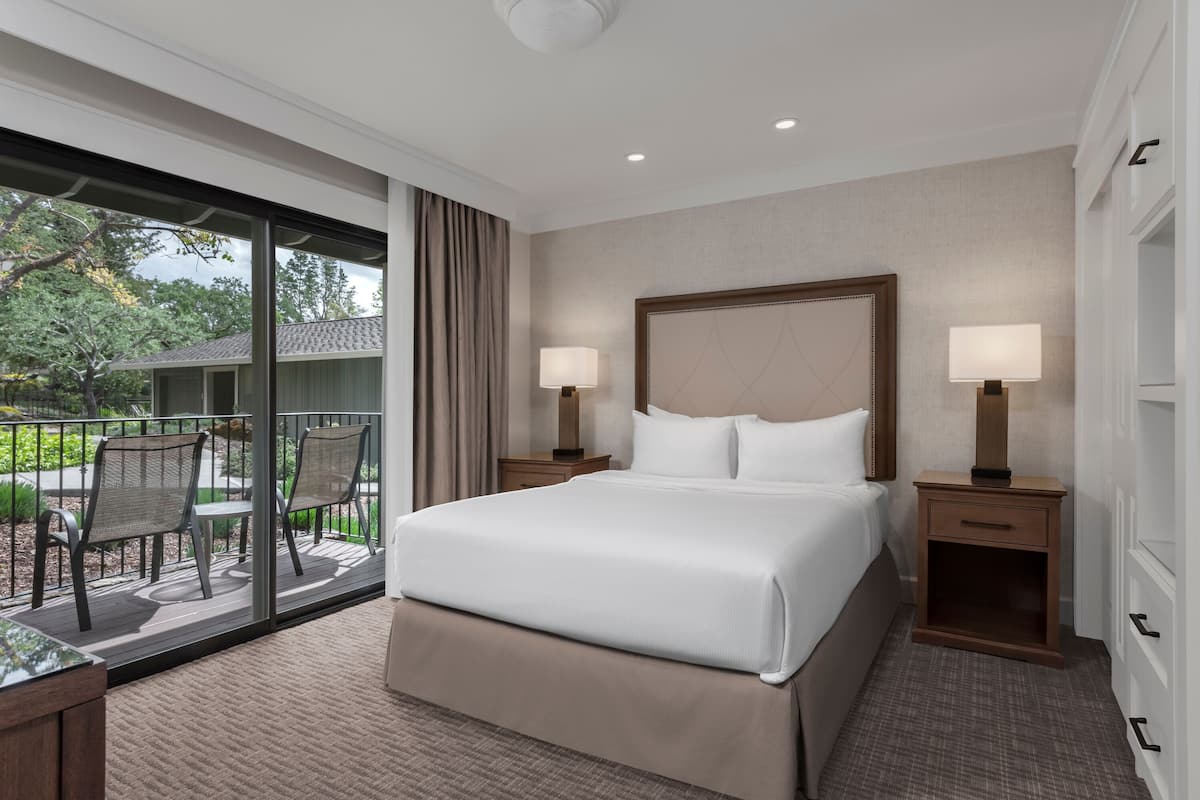 A hotel room with a large bed, two bedside tables with lamps, a sliding glass door to a balcony with outdoor furniture, and a view of trees and a building outside.