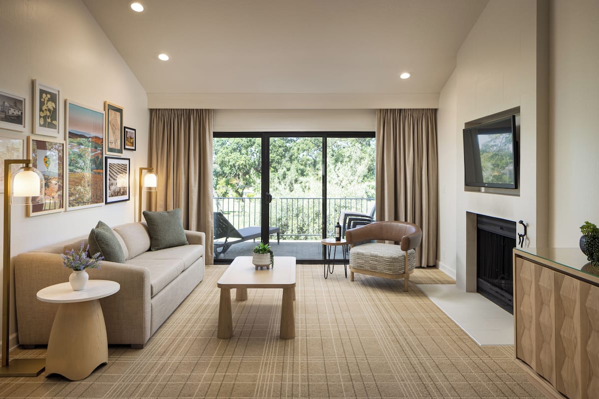 A living room with a sofa, a coffee table, a TV, a fireplace, and a balcony with a view of trees.