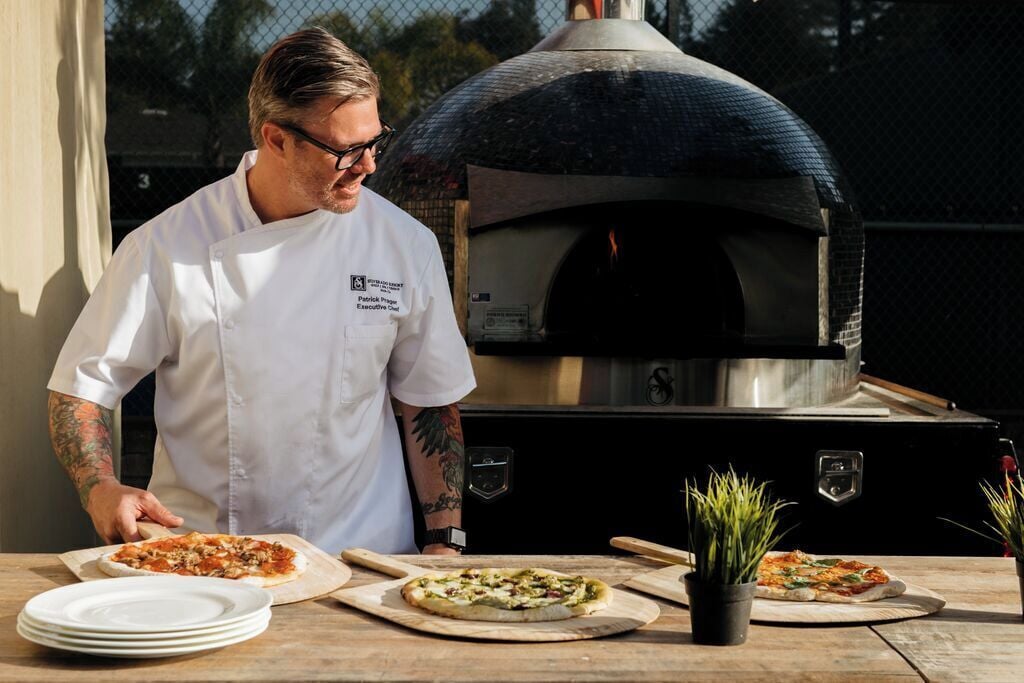 A chef in a white uniform with a name tag, preparing pizzas on wooden boards in front of a brick oven.