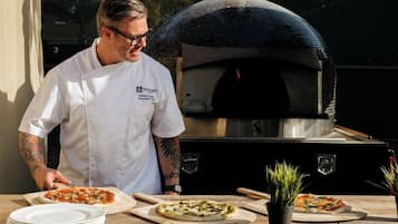 A chef in a white uniform with a name tag, preparing pizzas on wooden boards in front of a brick oven.