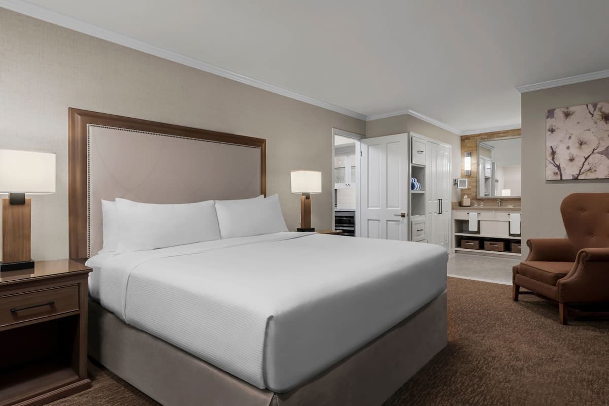 A hotel room with a large bed, bedside tables, a chair, a bathroom with a vanity, and a mirror.