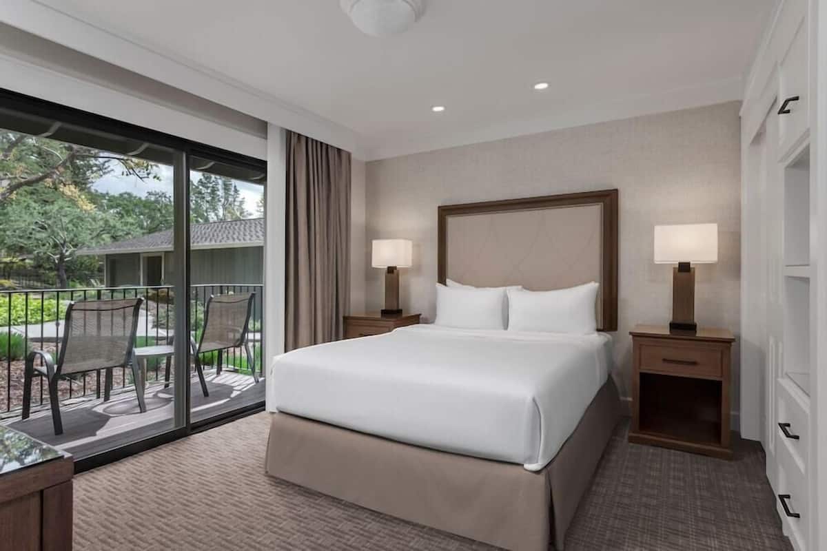 A hotel room with a large bed, two bedside tables with lamps, a sliding glass door to a balcony with chairs, and a view of trees and a building outside.