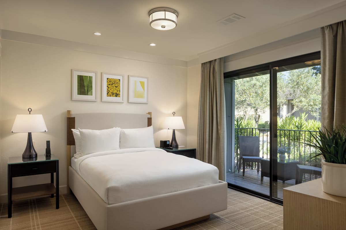 A hotel room with a large bed, two bedside tables with lamps, a sliding glass door to a balcony, and three framed pictures on the wall.