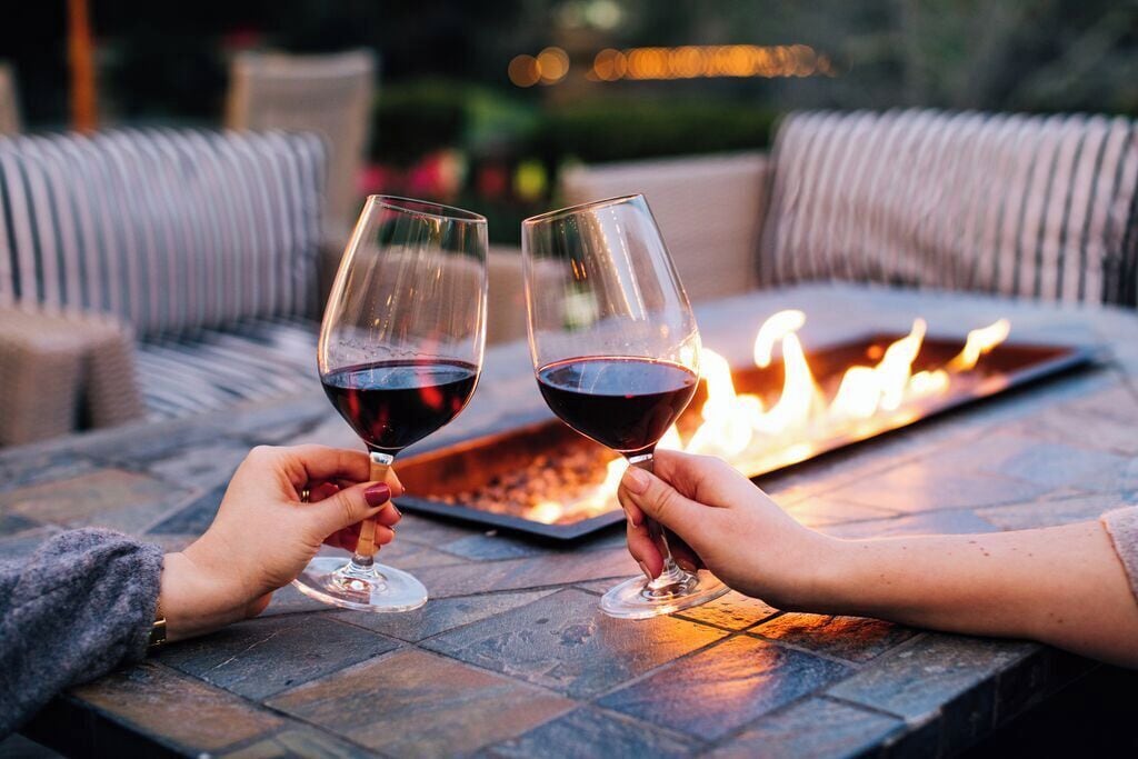 Dos personas brindando con copas de vino en una mesa al aire libre junto a una chimenea.